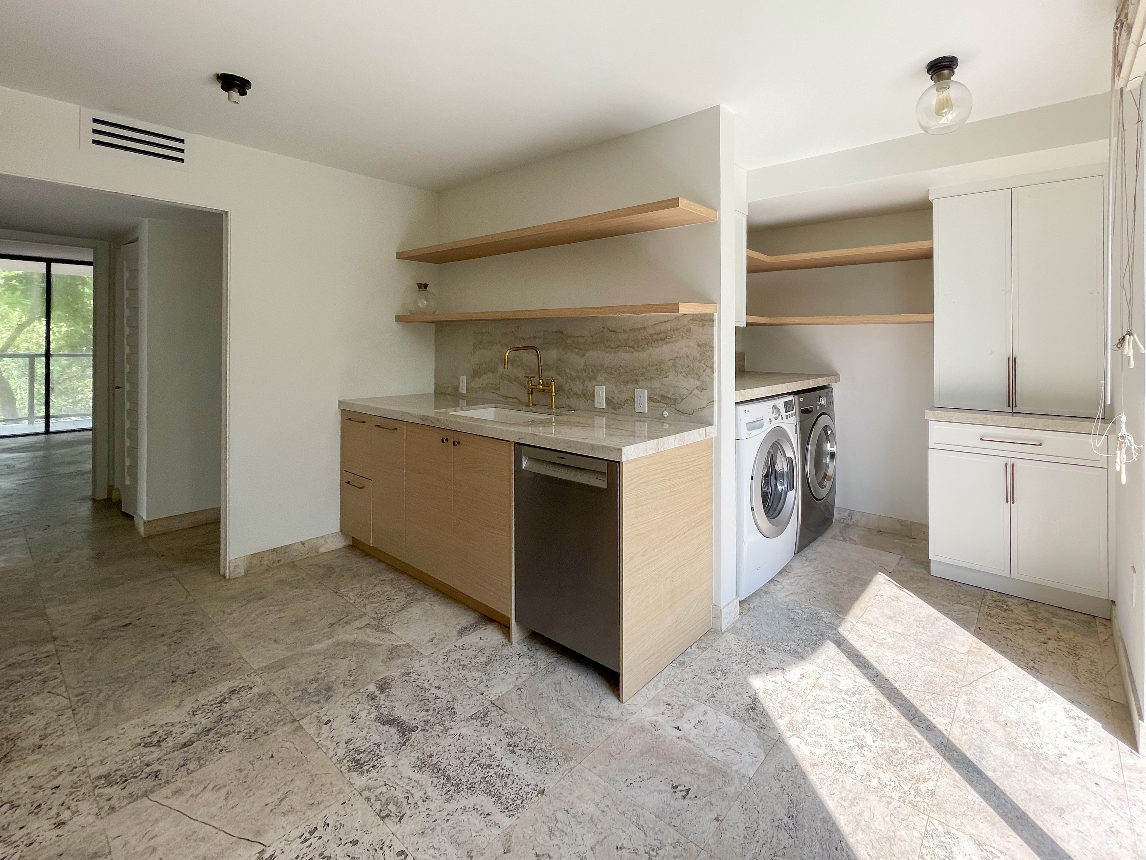 Open kitchen and laundry area with wood cabinetry, brass fixtures, and travertine floors